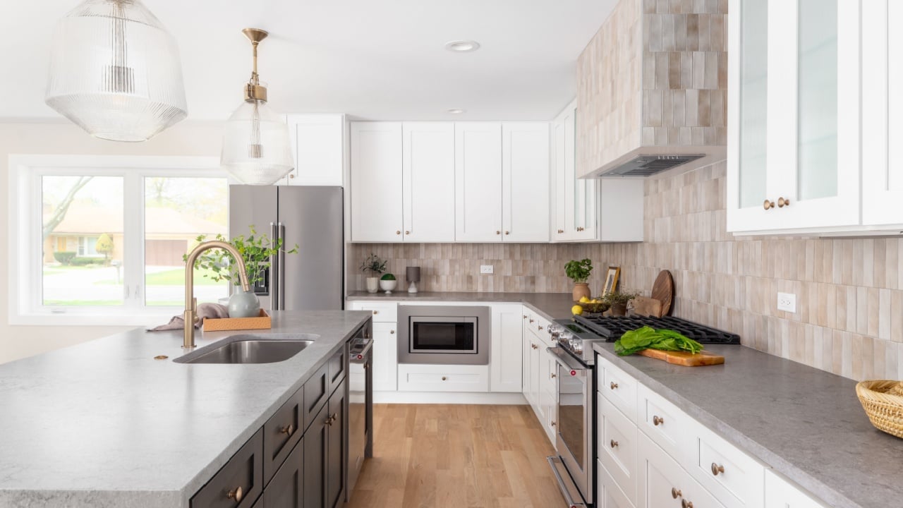 A white kitchen detail with stone countertops, gold light fixtures above the island, and a brown tiled backsplash. No brands or logos.