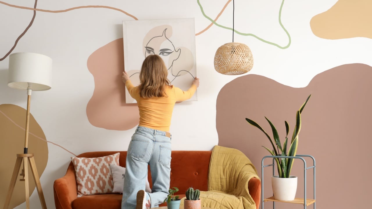 Young woman hanging stylish painting on wall at home