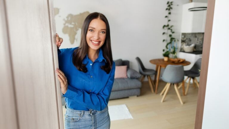 Joyful European lady opening door of her home and smiling to camera, woman meeting you in doors.