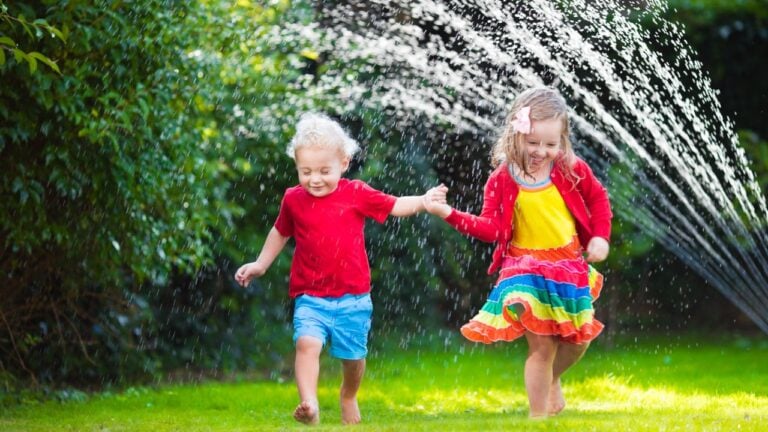 Child playing with garden sprinkler. Preschooler kid run and jump. Summer outdoor water fun in the backyard. Children play with hose watering flowers. Kids splash on sunny day.