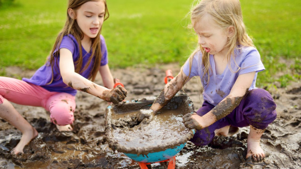 Two funny little girls playing in a large wet mud puddle on sunny summer day. Children getting dirty while digging in muddy soil. Messy games outdoors.