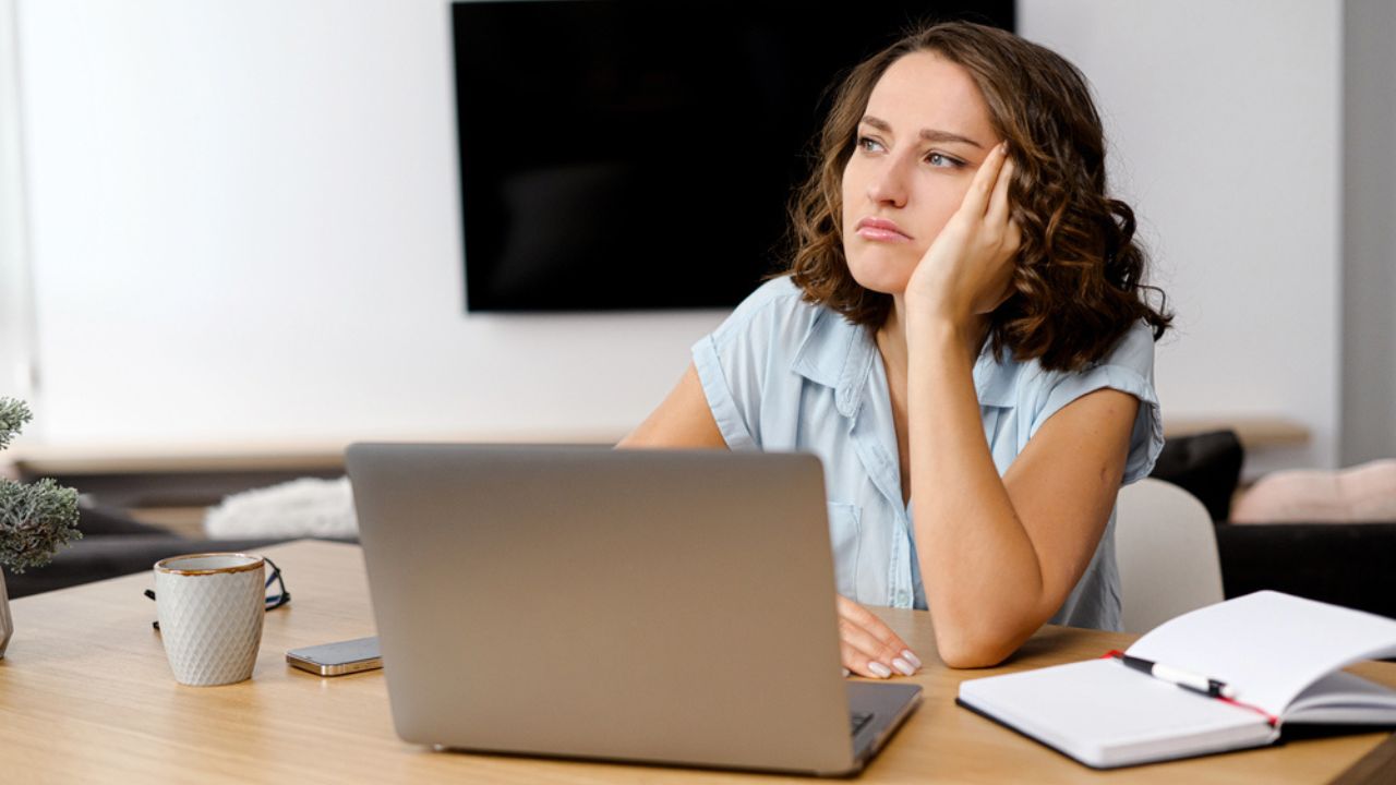 Young caucasian woman looking annoyed and stressed, sitting at the desk, using a laptop, thinking and looking away, feeling tired and bored