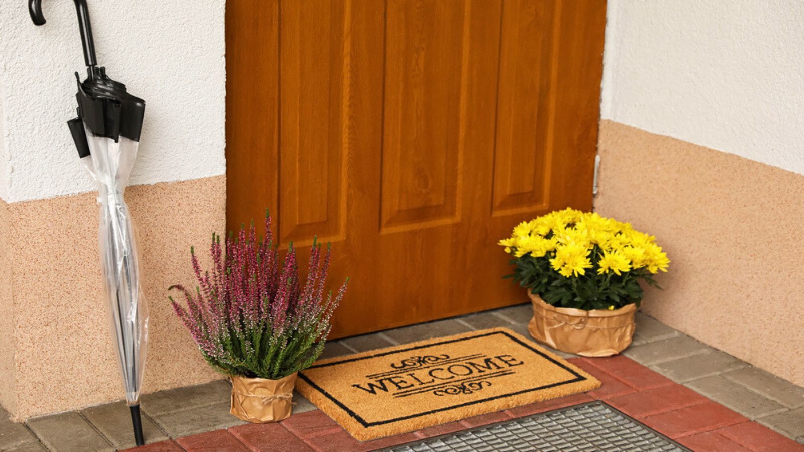 Door mat with word Welcome, umbrella and beautiful flowers near entrance