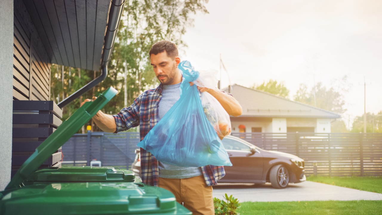 Caucasian Man is Throwing Away Two Plastic Bags of Trash next to His House. One Garbage Bag is Sorted with Biological Food Waste, Other with Recyclable Bottles Garbage Bin.