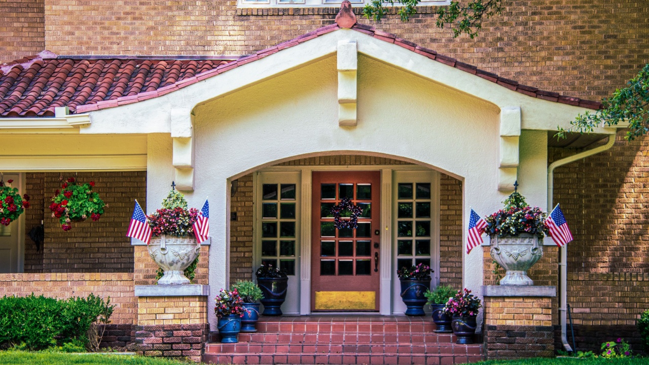 Entrance and stuco bungalow porch with french doors and tile roof on brick house with many flower pots and American flags and star shaped door wreath