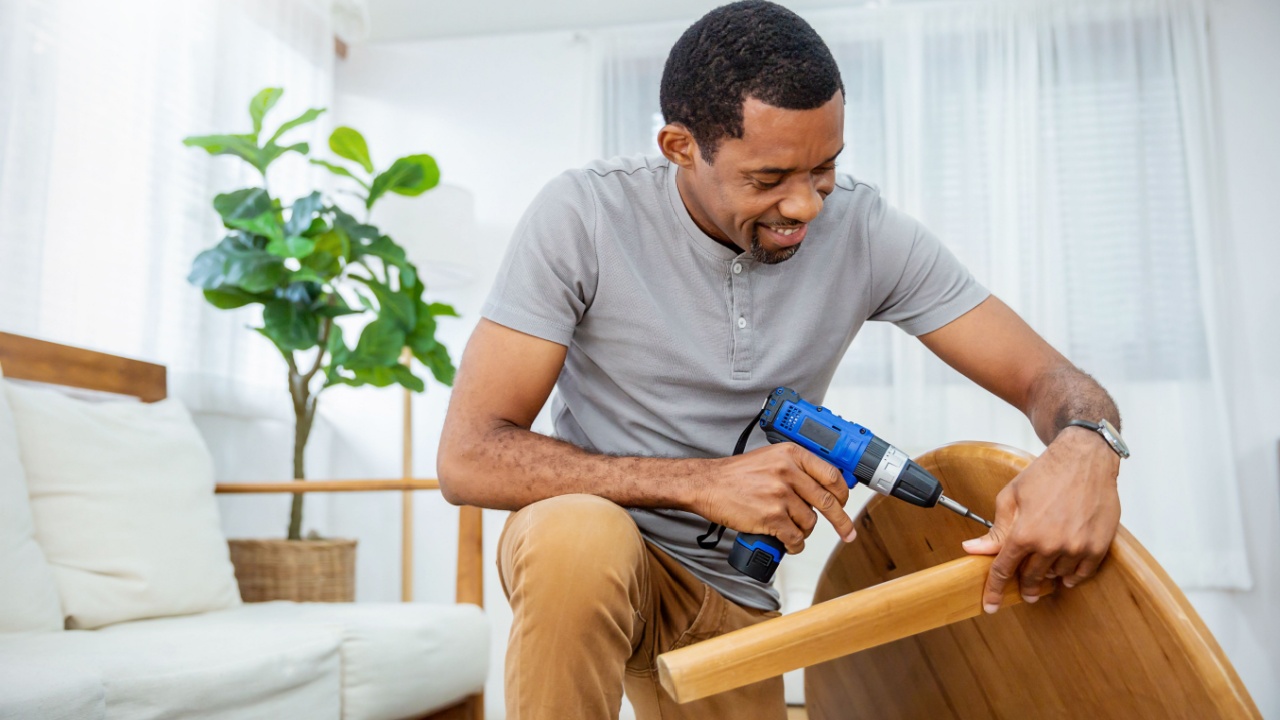 Portrait of happy busy African American black man screwing table with electric drill interior living room. DIY hand craft carpenter worker.