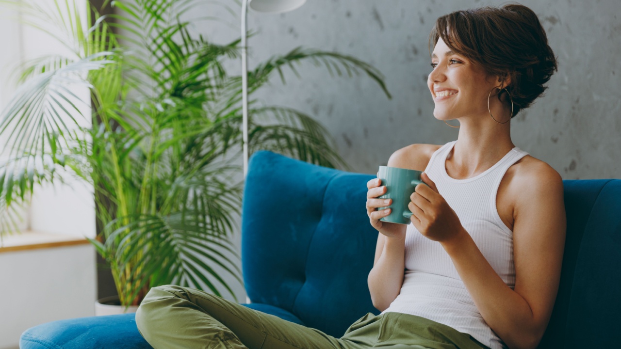 Side view young woman wears white tank shirt drink tea coffee sit on blue sofa couch stay at home hotel flat rest relax spend free spare time in living room indoors grey wall.