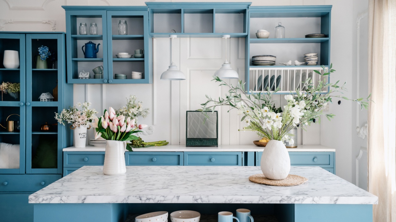 Marble countertop with vases and flowers in provence style apartment. Kitchen island and dining table with tableware. Blue furniture and white walls in classic interior design room.