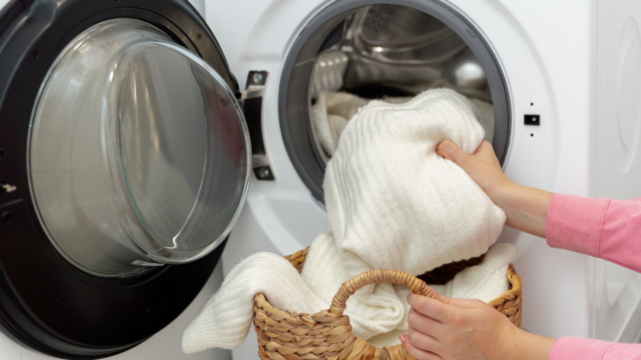 Woman putting white wool sweater and clothes into the drum of a washing machine in laundry room. Washing dirty clothes in the washer