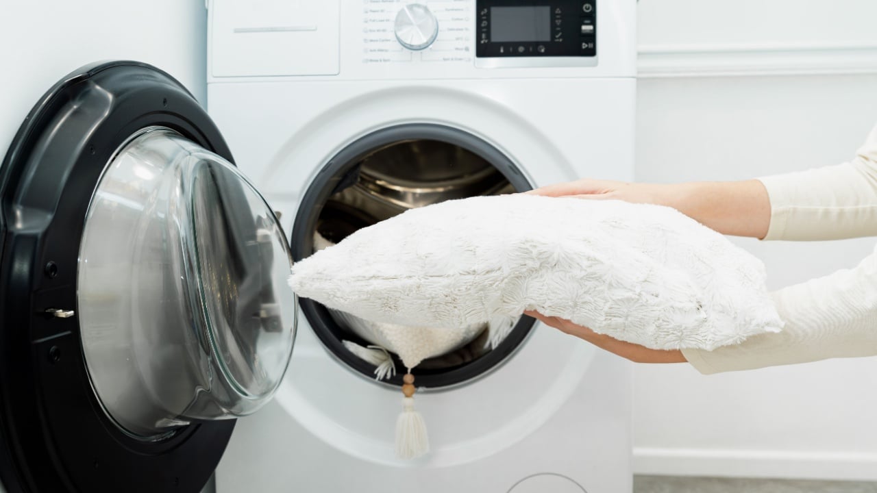 Woman holding clean white pillow in front of the drum of washing machine in laundry room. Washing pillows in the washer, housekeeping concept