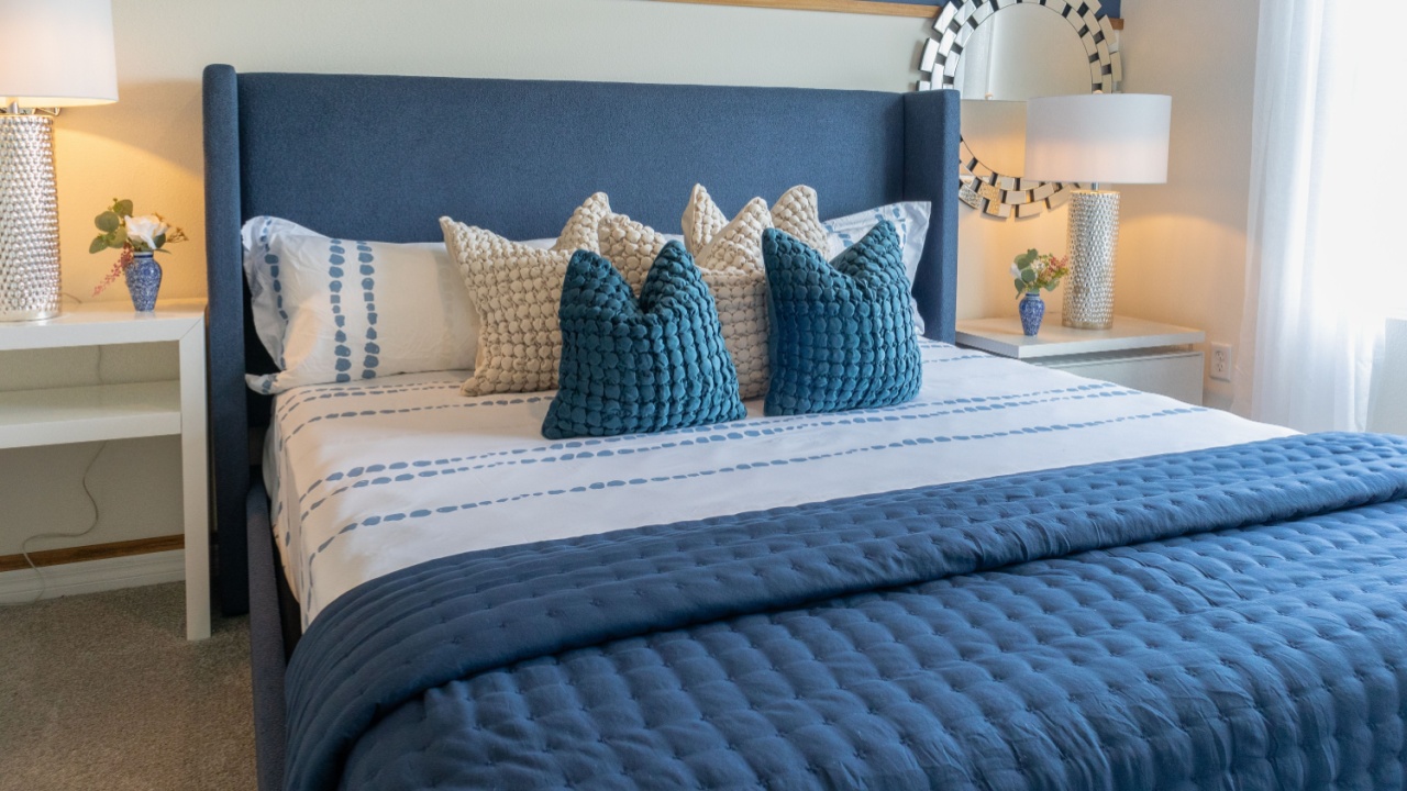 Front view of king size luxury bed with blue upholstered headboard in brightly lit master bedroom with two white night stands and silver lamps