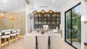 Modern kitchen interior with wooden cabinets, island seating, and large windows overlooking greenery.