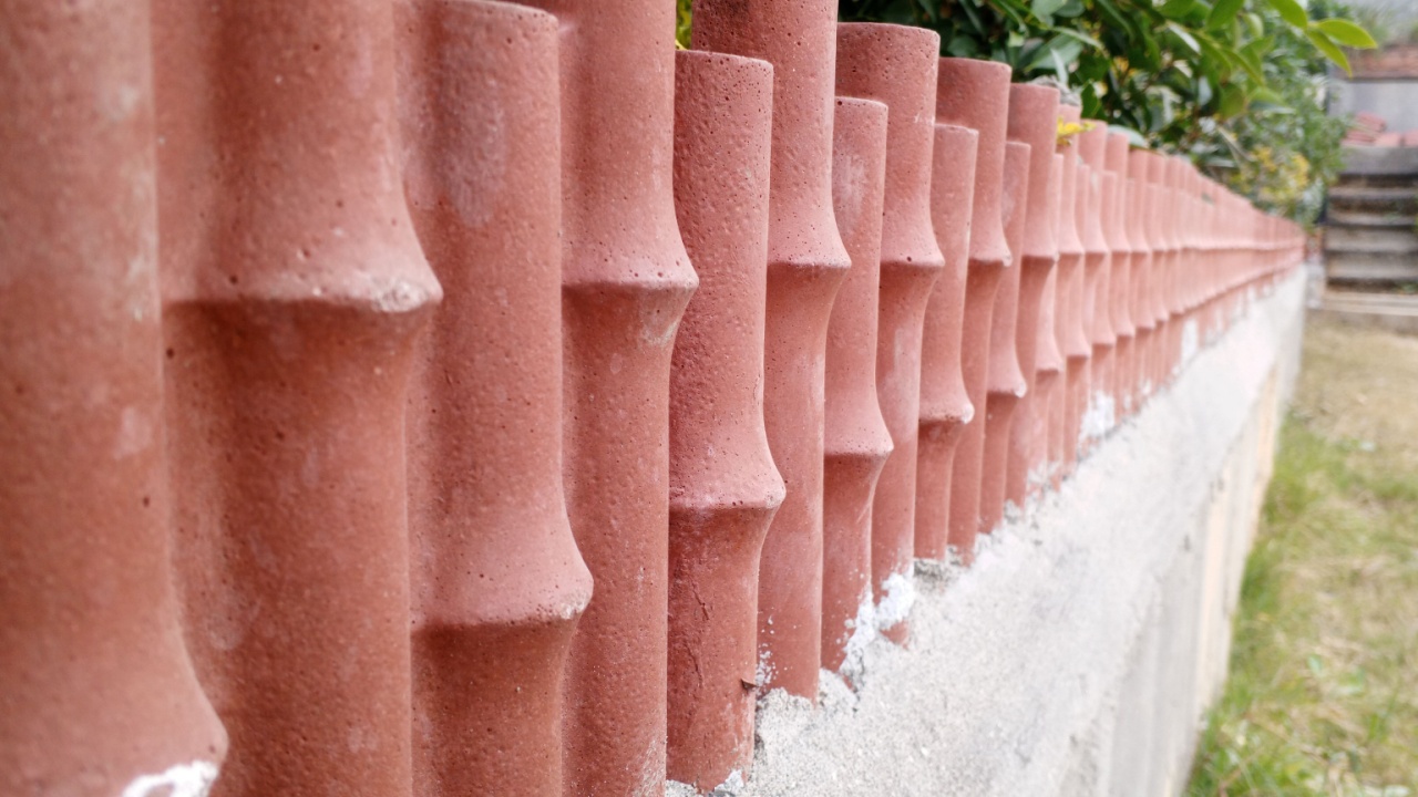 The image shows a boundary wall topped with decorative terracotta tiles shaped like bamboo stalks. The wall is made of concrete and has a rough, textured surface. Green plants peek over the wall from 