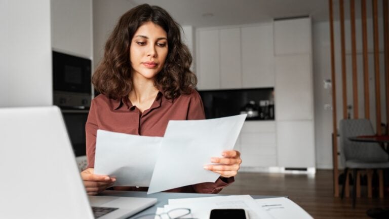 Focused latin hispanic young business woman working on laptop computer reading financial document report at home office. Accountant entrepreneur manager businesswoman doing paperwork using pc remotely