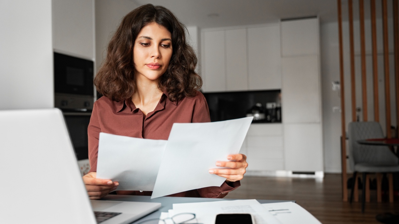 Focused latin hispanic young business woman working on laptop computer reading financial document report at home office. Accountant entrepreneur manager businesswoman doing paperwork using pc remotely