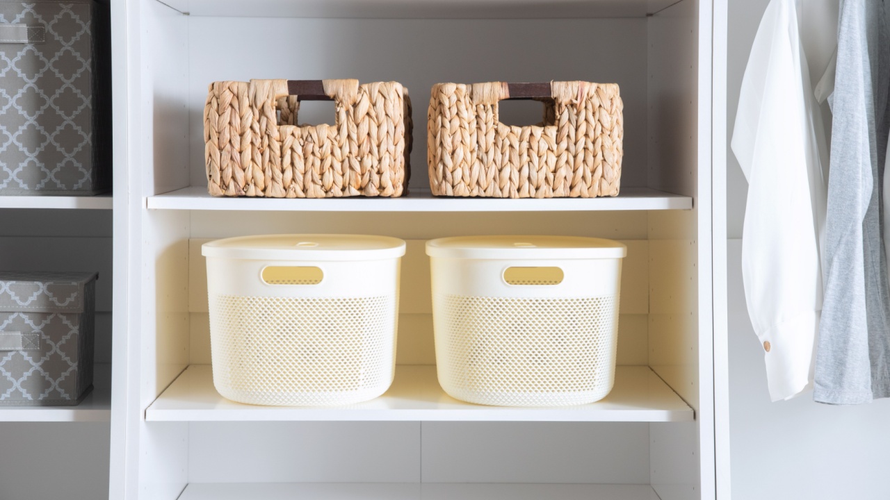 Scandinavian inspired closet shelf detail with woven baskets and cream plastic containers on white shelving, near gray patterned boxes and neutral clothing in a clean minimalist wardrobe setup.