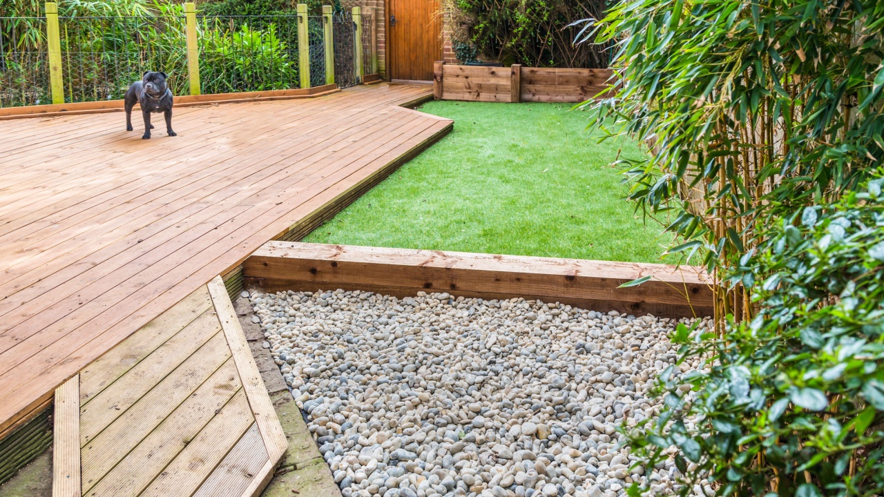 A section of a residential garden, yard with wooden decking, patio over a fish pond, a section of artificial grass and an area of stone pebble. There is a bamboo plant and a dog in the garden.