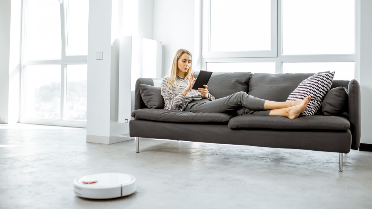 Woman relaxing on the couch while automatic vacuum cleaner doing the housework in the modern white living room.