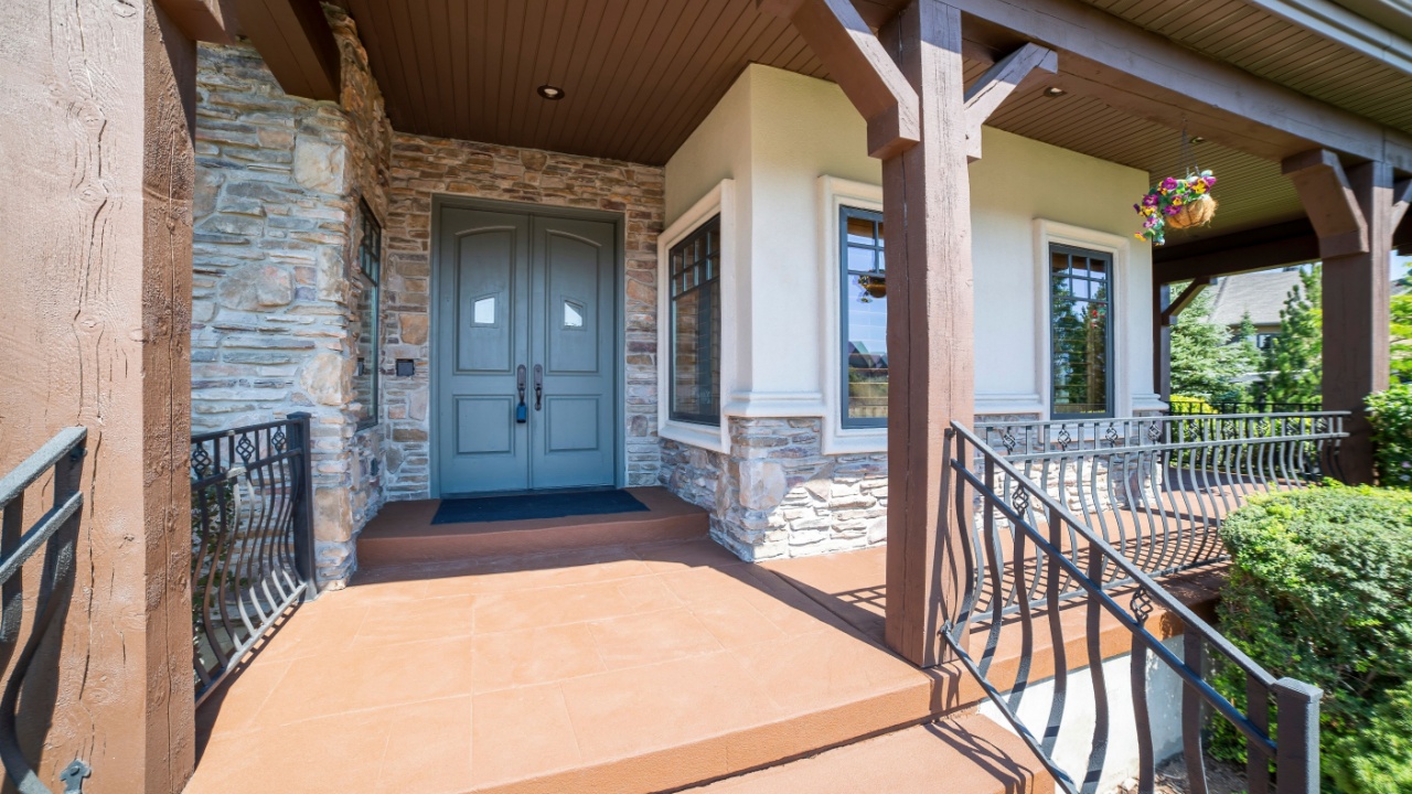 Porch with gray double entry doors beside the windows and stone walls. Entrance of a house with wooden column posts and stairs with stylish metal railings along with the bushes at the front.