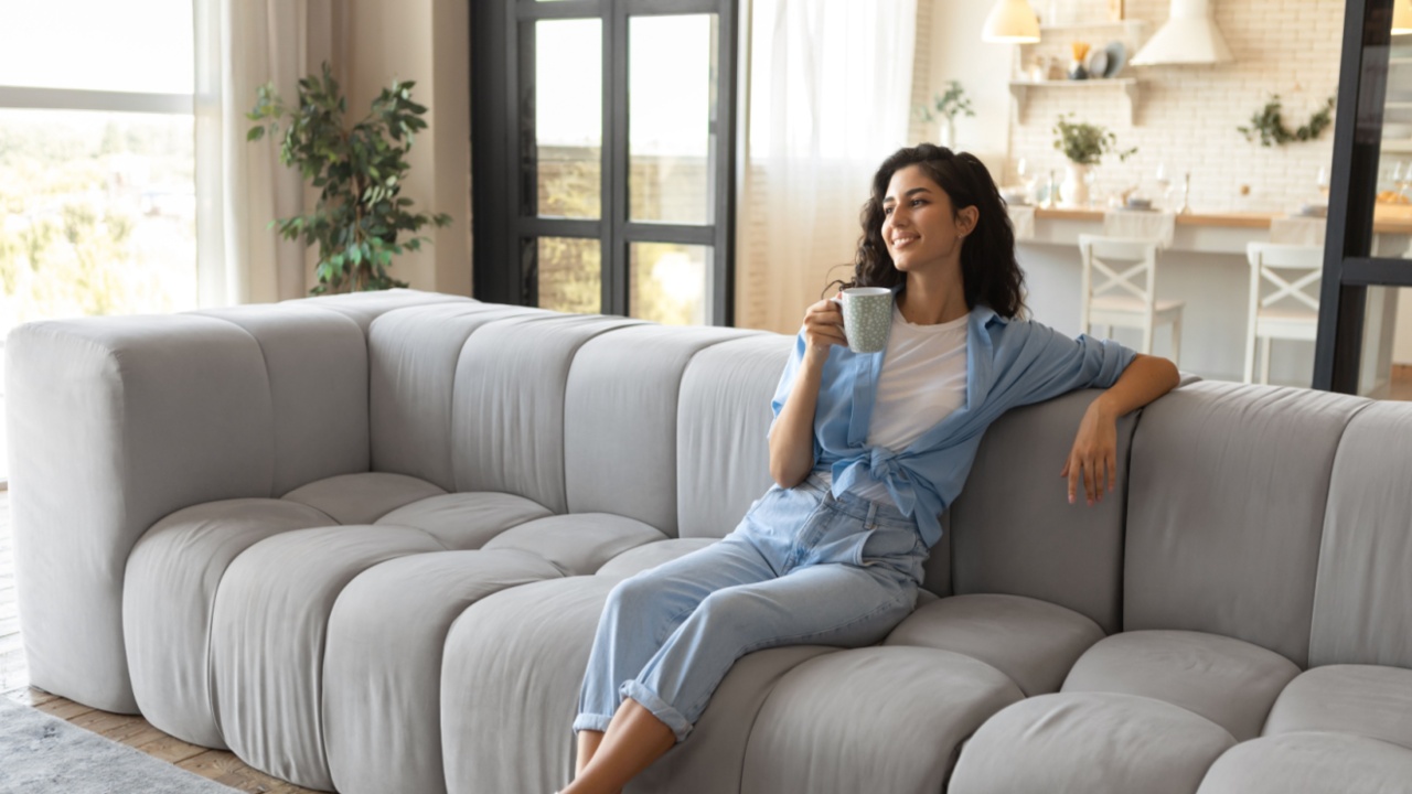 Attractive Caucasian woman enjoying lazy morning with coffee in living room, copy space. Beautiful young brunette lady relaxing on couch with cup of hot drink at home, full length