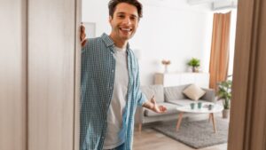Portrait of cheerful man welcoming inviting visitor to enter his home, happy young guy standing in doorway of modern apartment, millennial male holding door looking out showing living room with hand