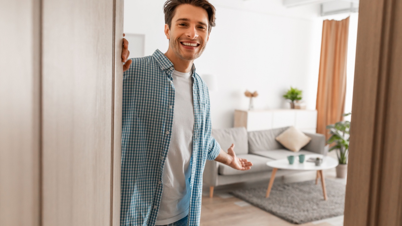 Portrait of cheerful man welcoming inviting visitor to enter his home, happy young guy standing in doorway of modern apartment, millennial male holding door looking out showing living room with hand