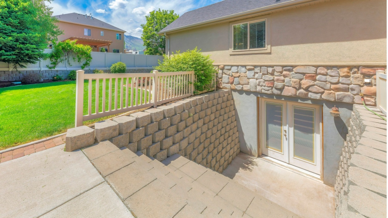 Panorama white clouds Basement entrance with glass panels on the double doors. There is a stairs near the stack of concrete blocks near the lawn and a view of bushes and trees near the fence