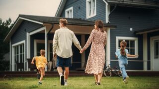 Young Beautiful Couple Walking Outdoors Towards the Country House, Holding Hands. Their Little Son and Daughter Running Together with Them. Young Modern Family at Home, Footage from The Back.