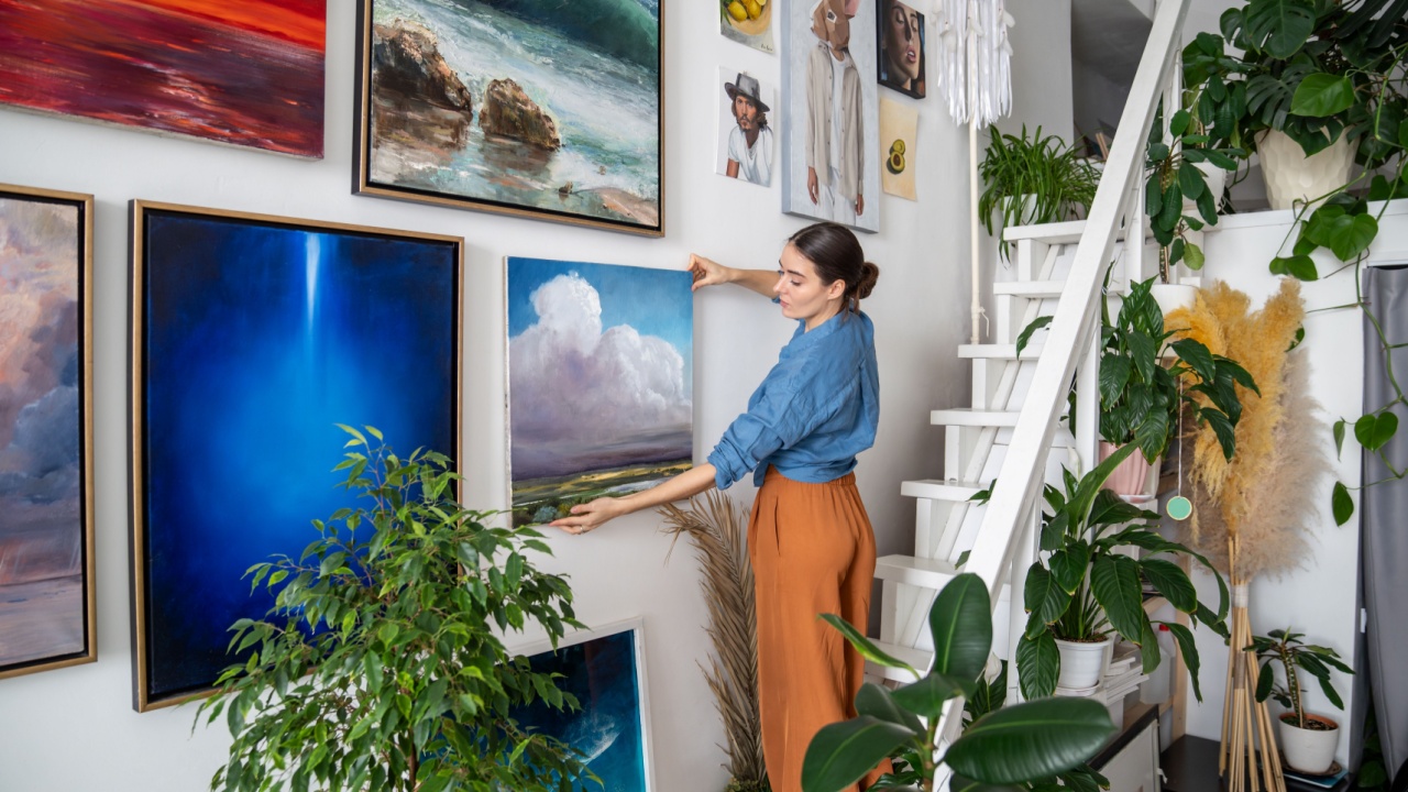 Serious woman hangs pictures on wall. Concentrated unsmiling female artist creates composition of paintings, decorations on wall. Creative designer in art home studio workshop among artworks plants.