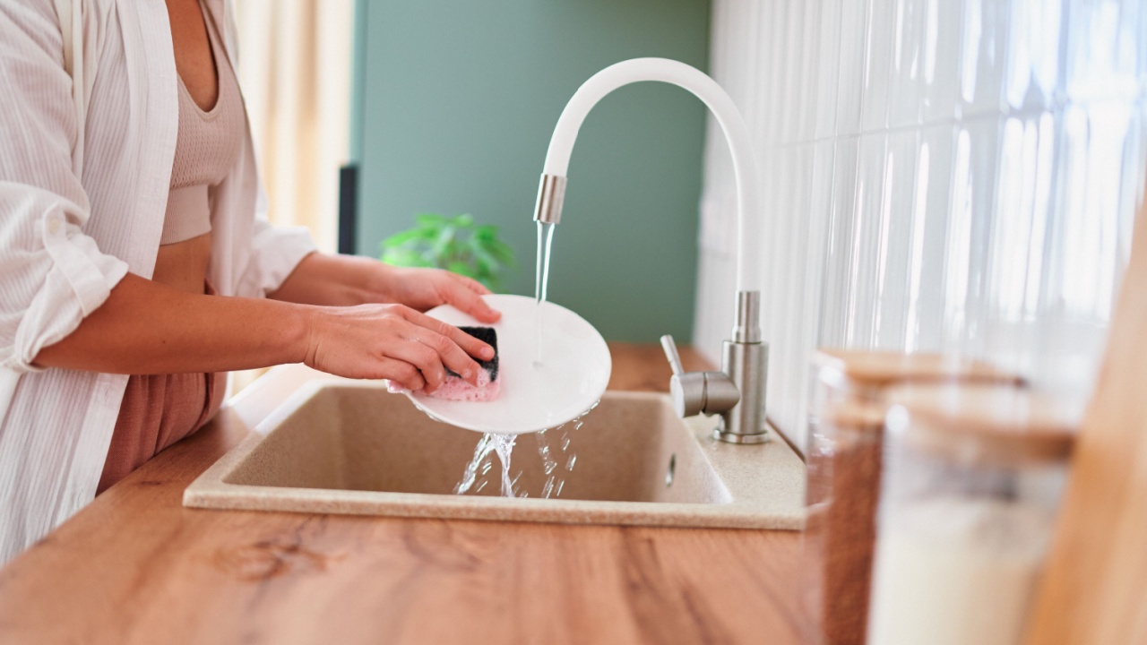 Woman washing dishes under running water from white tap using detergent and sponge in scandinavian style kitchen at home