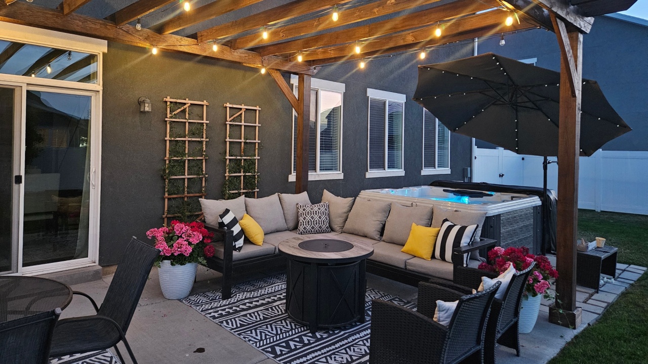 An outdoor patio with a grey L-shaped couch, striped and yellow pillows, black and white rugs, potted flowers, hot tub, fire pit, and extra black seating outside a house, creating an inviting space.