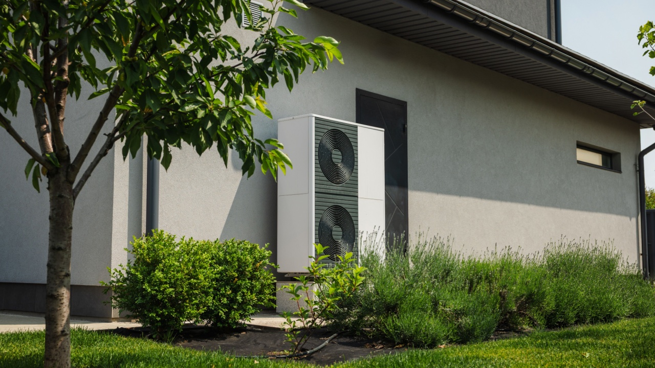 Modern heat pump unit installed next to a residential house wall, surrounded by green lawn, bushes, and young trees in a landscaped yard.