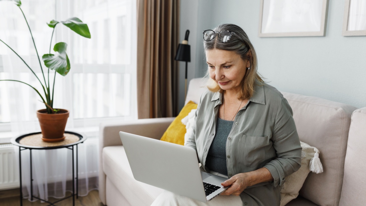 Side view indoor image of aged woman in casual clothes using laptop or study online at home, sitting in cozy tidy living-room on couch and browsing social media, surfing internet, reading news