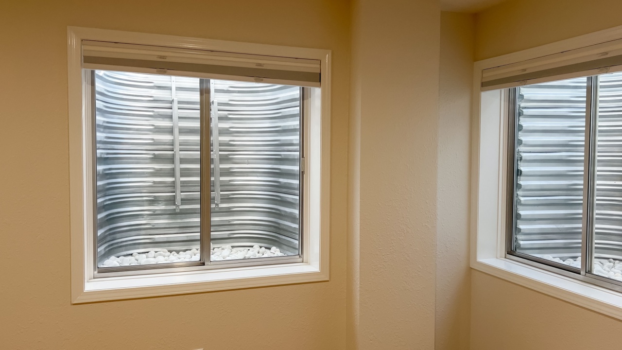 A centered view of a single egress window in a beige-painted basement room. The window is framed with white trim and looks out onto a corrugated metal well filled with white rocks.