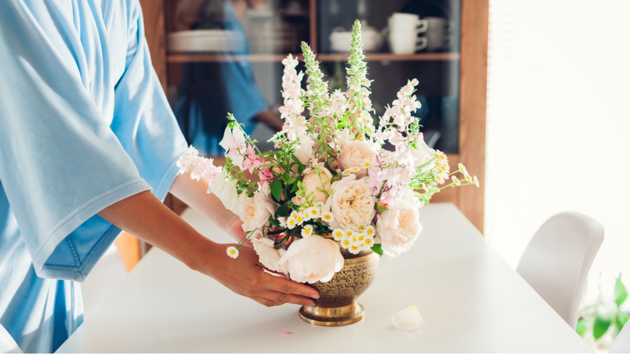 Woman puts brass vase with bouquet full of roses flowers on table by cupboard on kitchen. Interior and home decor. Summer floral arrangement.
