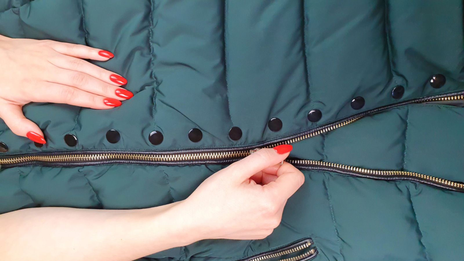 Female hands with red nails close zipper lock on down jacket. Autumn jacket fabric background, green puffer jacket texture. Top view
