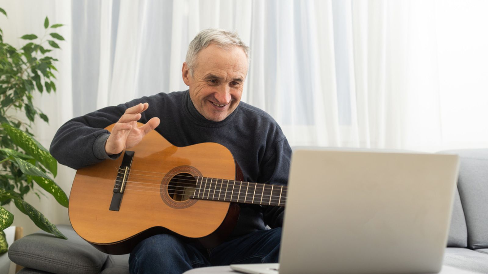 Portrait of senior man in headphones taking online guitar lesson looking at laptop screen. Retired male learning to play guitar watching webinar on computer at home