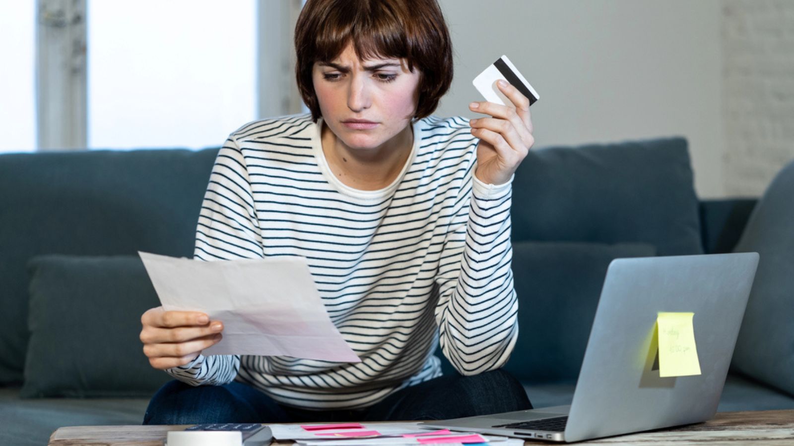Portrait of worried and desperate woman feeling stressed paying credit card debts and bills on laptop computer on sofa at home. In business and home finances and financial problems concept