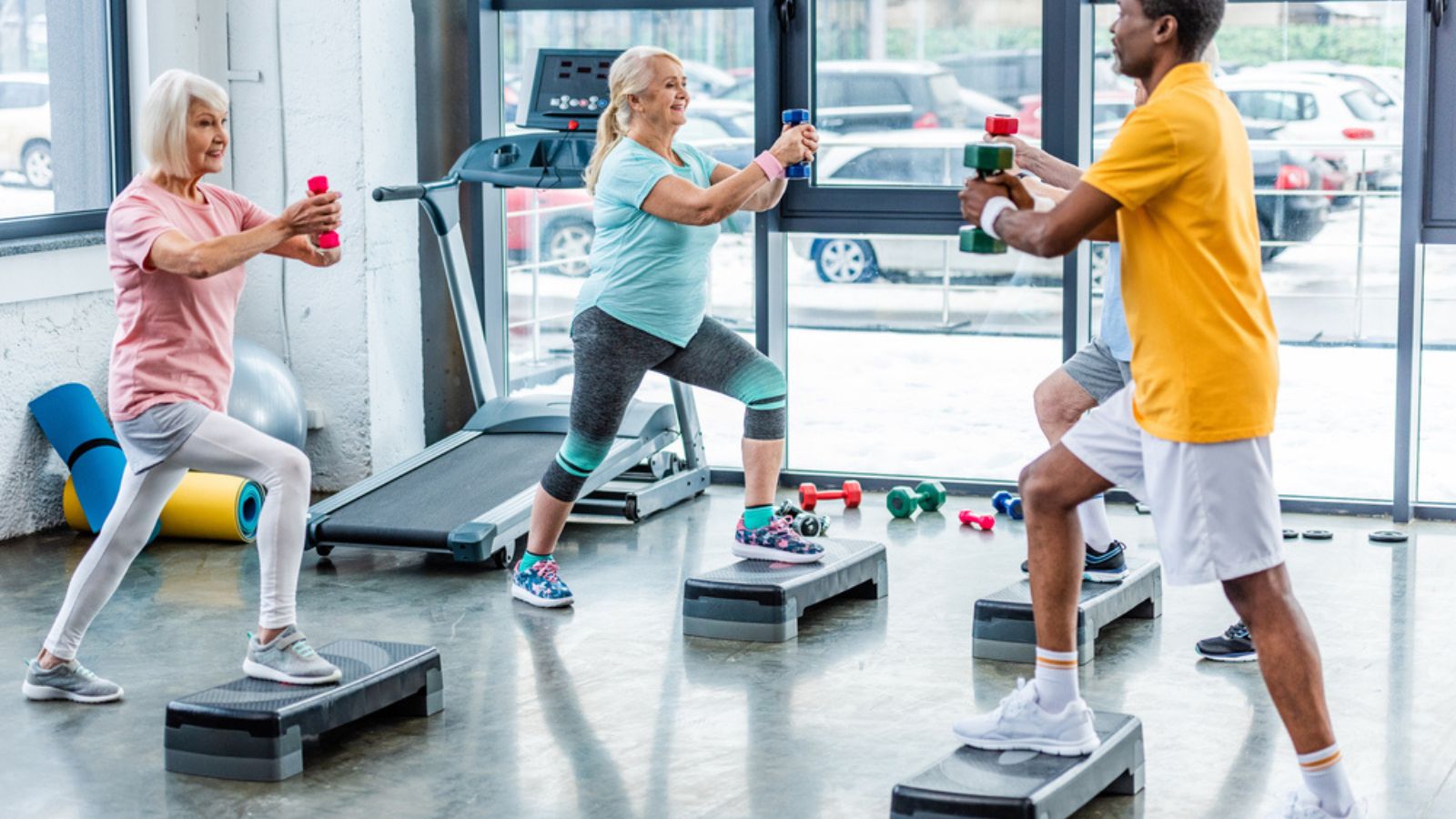 Selective focus of senior multicultural sportspeople synchronous exercising with dumbbells on step platforms at gym