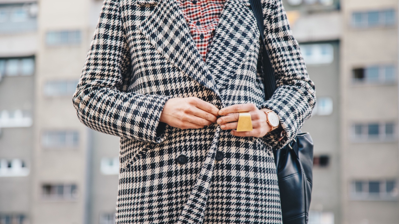 detail shot of a woman outdoors posing in a checked patterned coat, wearing a golden ring and watch and holding a black backpack. fashion outfit details, lady buttoning up her coat