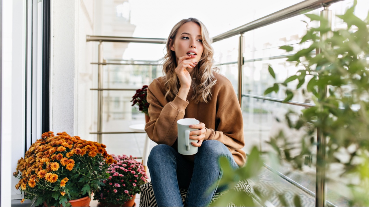 Romantic girl sitting near flowers with cup of coffee. Carefree young woman thinking about something.