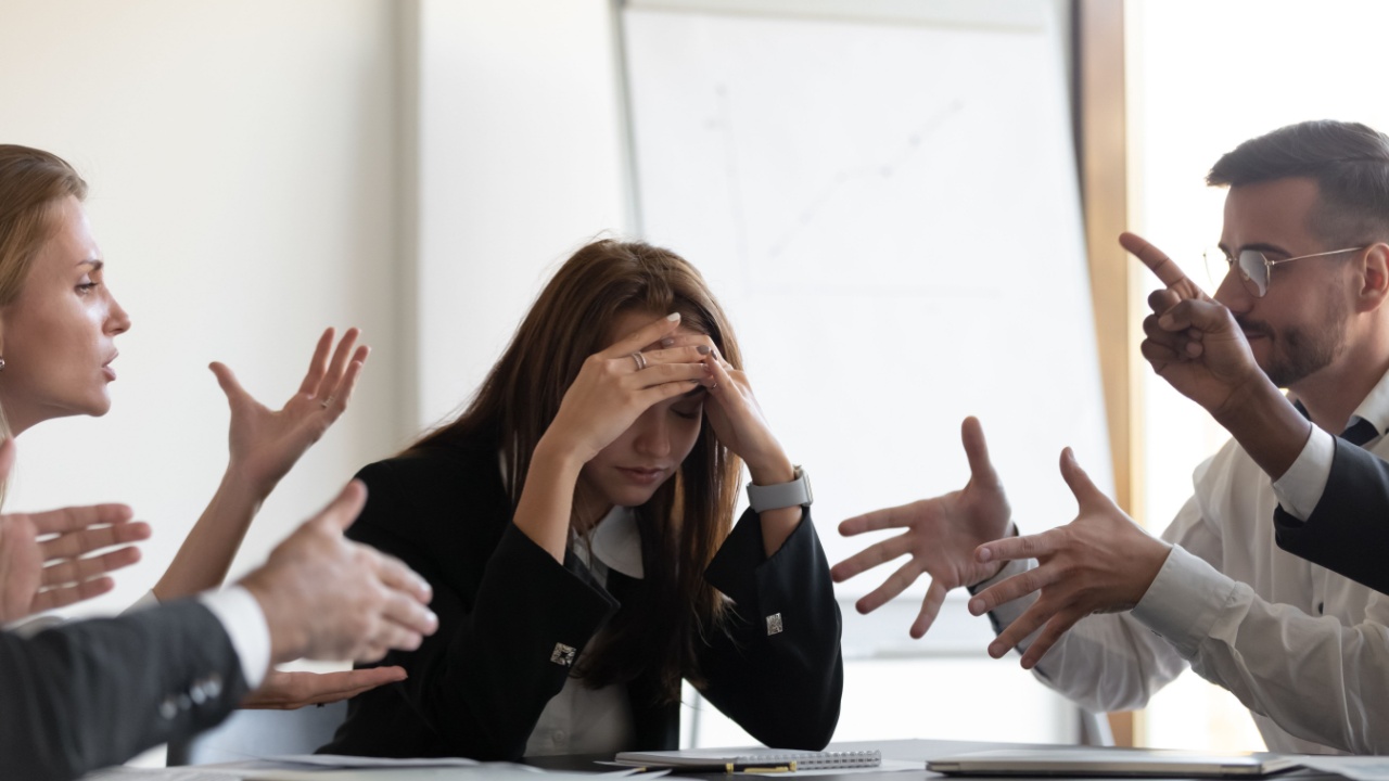 Frustrated millennial female worker sitting at table with colleagues, felling tired of working quarreling at business meeting. Upset stressed young businesswoman suffering from head ache at office.