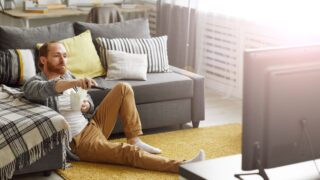 Full length portrait of bearded man switching channels while watching TV at home in bachelors pad, copy space