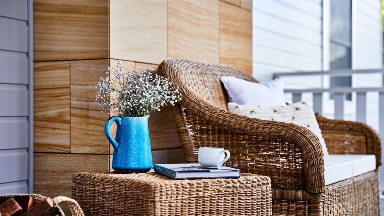 Photography of a cane furniture setting on the front porch of a grey timber home with a sandstone detail, a blue jug of flowers, books and a cup of tea