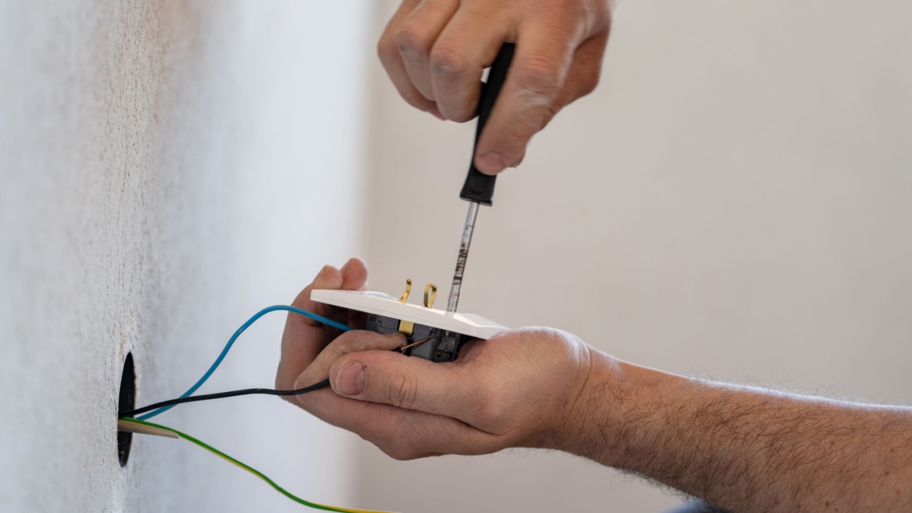 Close up on hands of caucasian man electrician holding screwdriver working on the plug electric on residential electric system installing white AC power socket on gray wall at home repair close up