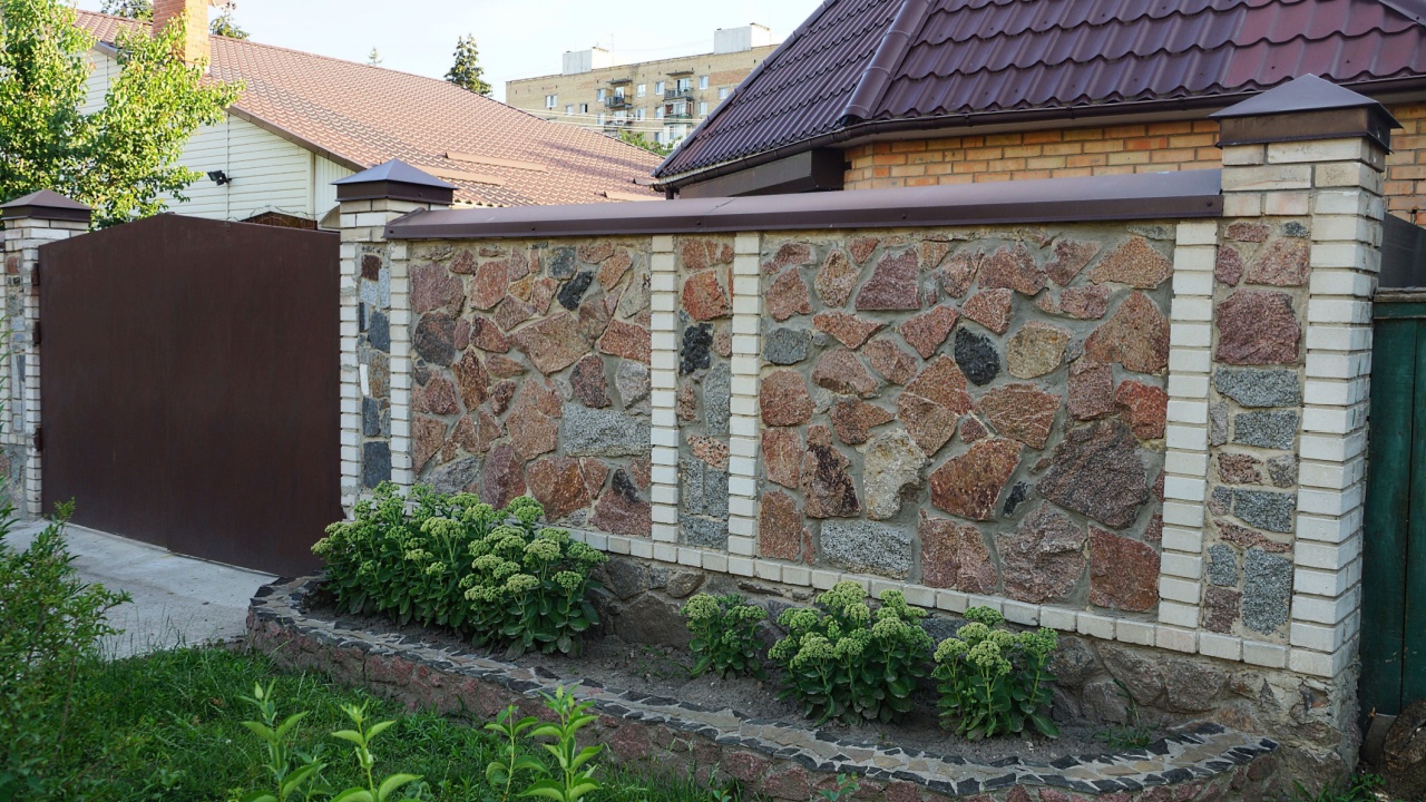 part of a brown stone wall of a fence from a closed metal gate in green grass and vegetation on the street