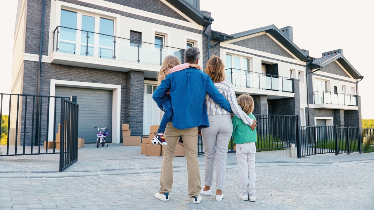 Back view of happy family with small children standing near their new modern house and hugging at suburb and looking on it.