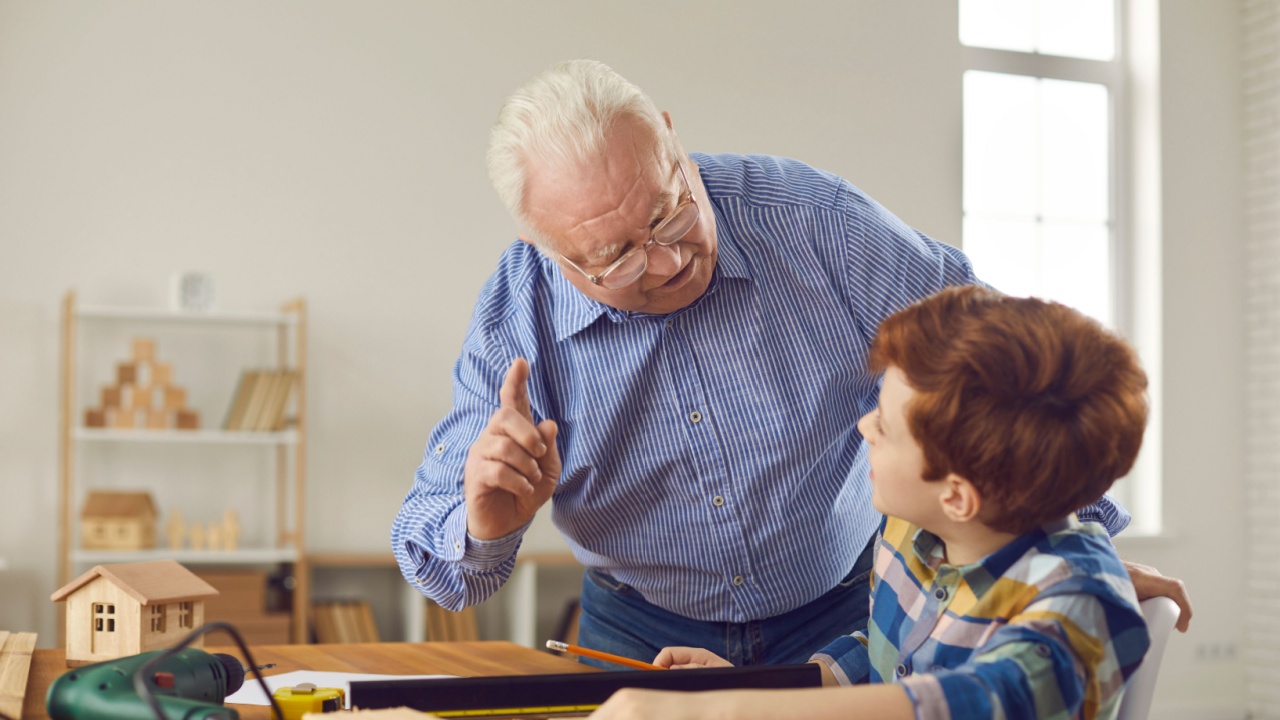 Wise senior carpenter reminds little child of safety precautions when working with wood at the workshop. Grandfather in retirement gives grandson useful advice on how to use repair tools at home