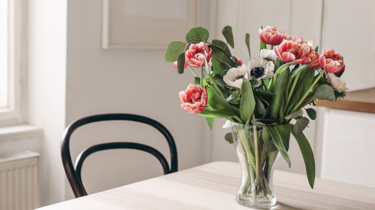 Spring flowers in glass vase on wooden table. Blurred kitchen background with old chair. Bouquet of red tulips, white anemone flowers and eucalyptus branches. Contemporary elegant scandi interior.
