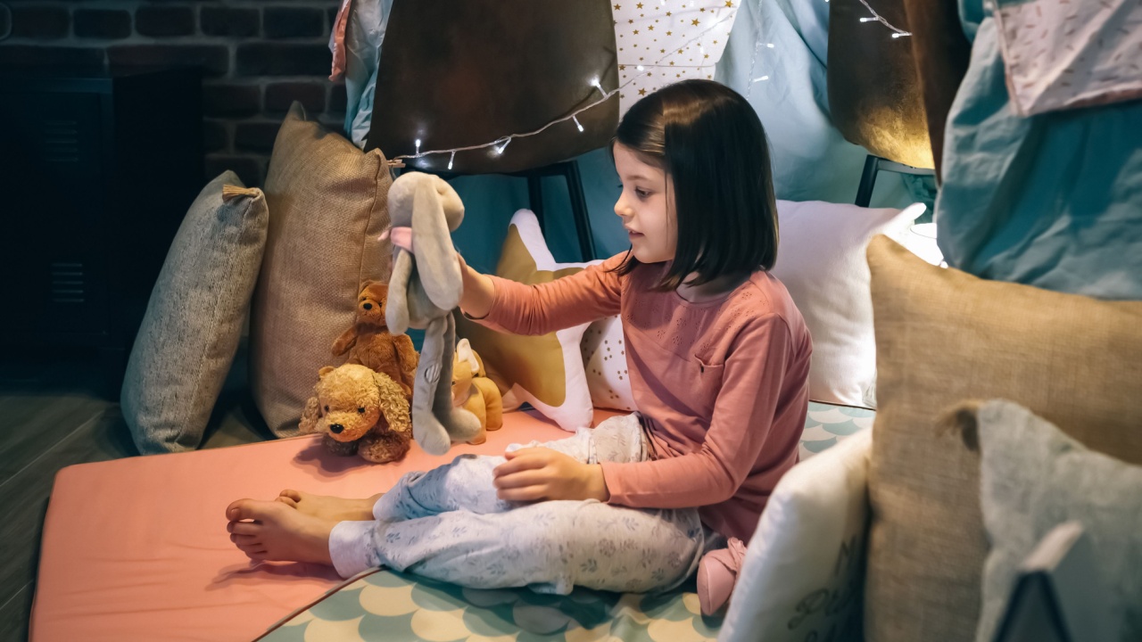 Adorable girl in pajamas playing alone with teddies in a play shelter made of bed sheets and chairs